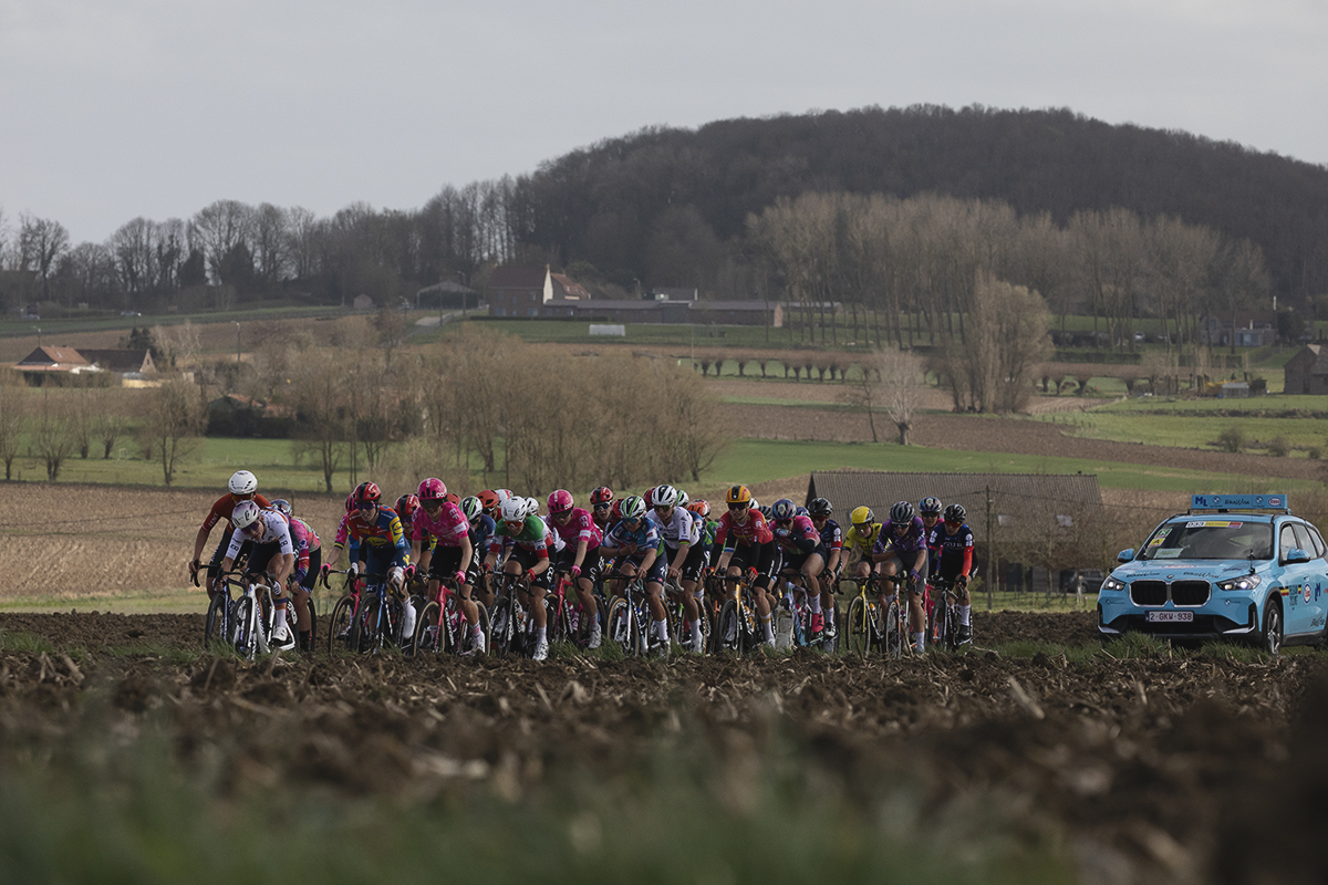 Gent Wevelgem Vrouwen 2025 - Riders seen over a ploughed field with the rise of a distant tree lined hill behind