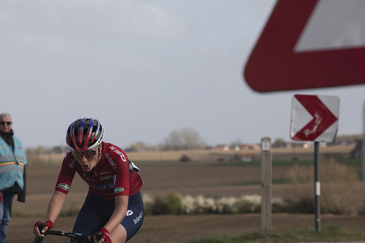 Gent Wevelgem Vrouwen 2025 - Kaja Rysz rides round a bend with the red chevron and red of a road sign matching her kit