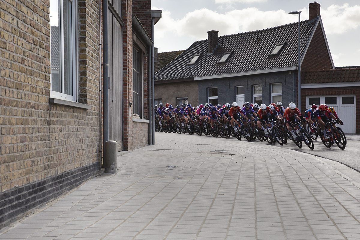 Gent Wevelgem Vrouwen 2025 - The peloton rounds a corner in Reningelst past typically Belgian buildings