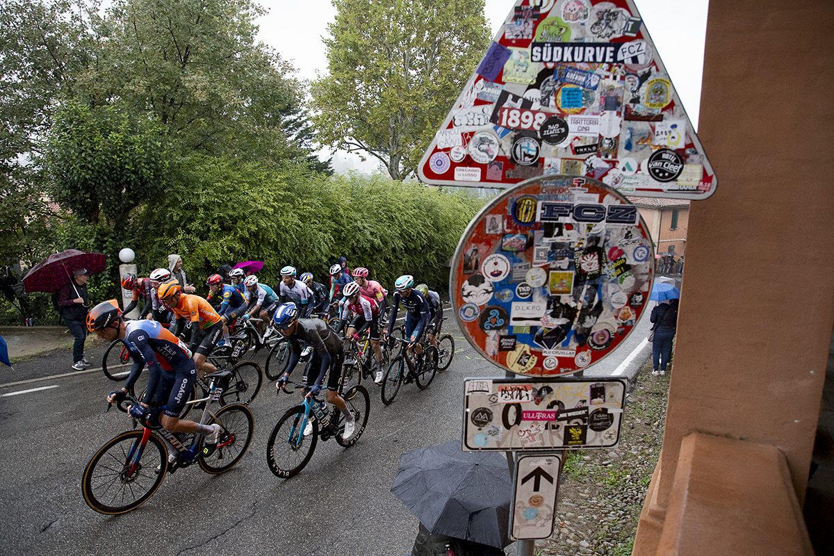 Giro dell’Emilia 2024 - Riders pass a sign covered in stickers on Via di San Luca