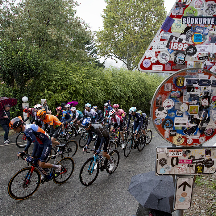 Giro dell’Emilia 2024 - Riders pass a sign covered in stickers on Via di San Luca