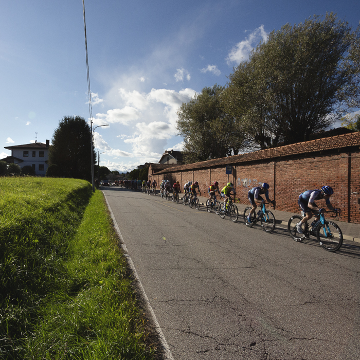 Gran Piemonte 2024 - A line of riders race into Maggiora with a wall to one side and open fields to the other