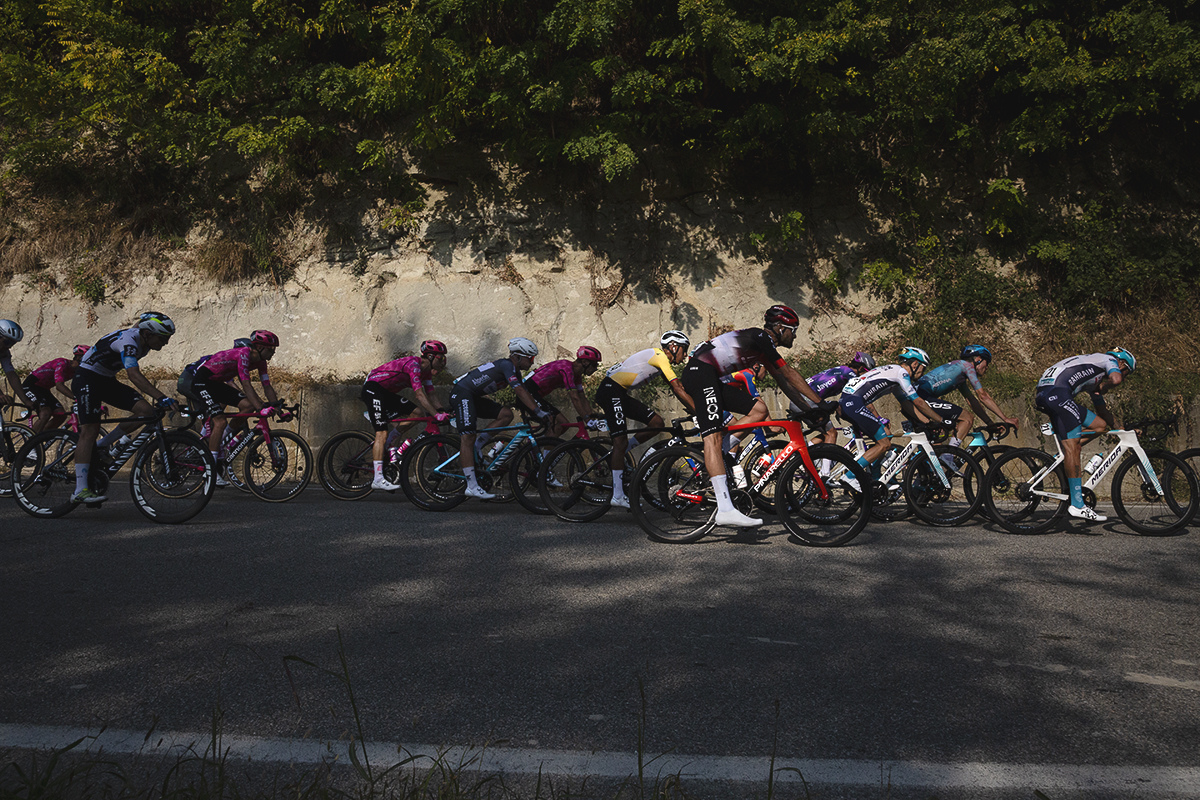 Gran Piemonte 2025 - The peloton is lit by the afternoon sunlight as it passes by a honey coloured rock face near Castel Boglione