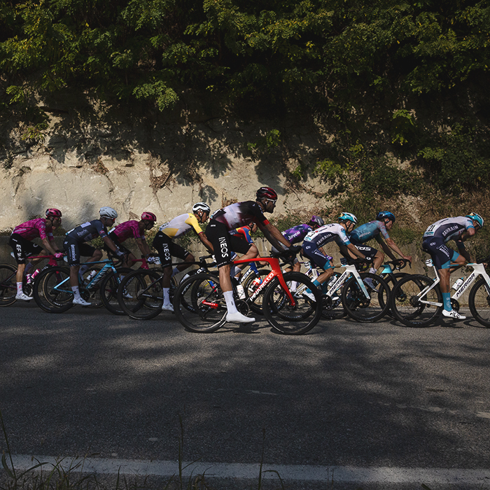 Gran Piemonte 2025 - The peloton is lit by the afternoon sunlight as it passes by a honey coloured rock face near Castel Boglione