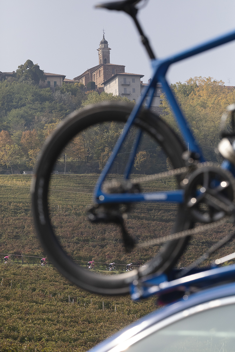 Gran Piemonte 2025 - Riders seen from a distance framed by a bike wheel on top of one of the race convoy