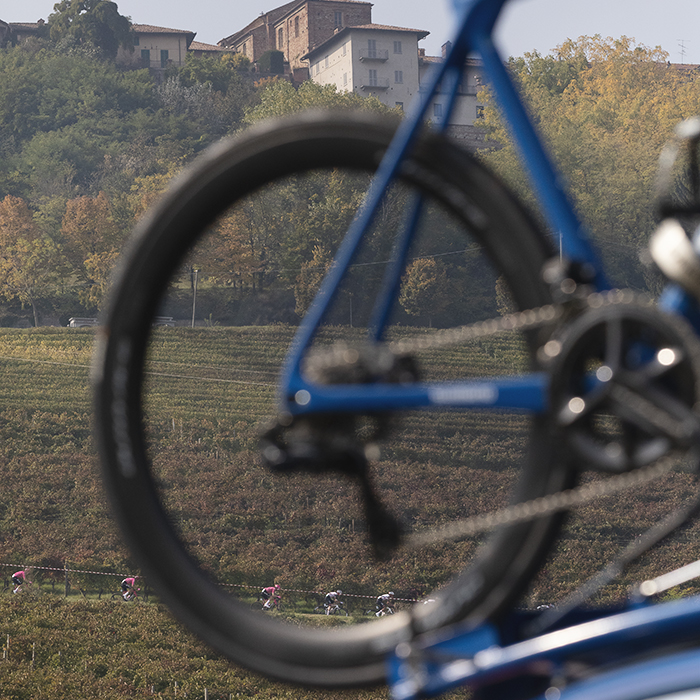 Gran Piemonte 2025 - Riders seen from a distance framed by a bike wheel on top of one of the race convoy
