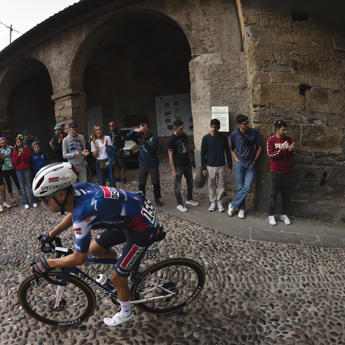 Il Lombardia 2025 - Junior Lecerf rounds a corner on the cobbles as fans stand in a small portico behind