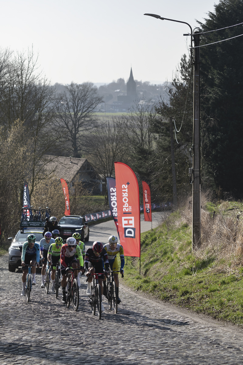 Kuurne Brussel Kuurne 2025 - A group of riders climbs the cobbles of Mont St-Laurent with the countryside in the background