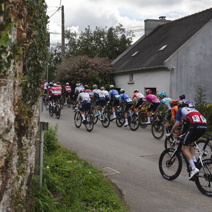 Bretagne Classic - Ouest-France 2025 - The peloton moves away down the road framed by a stone wall and small cottage