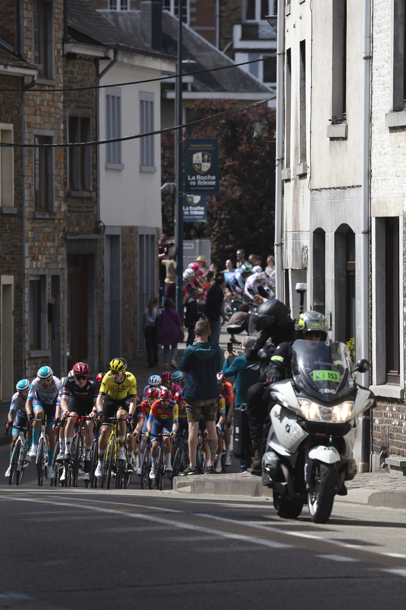 Liege Bastogne Liege 2025 - The peloton approaches up a road lined with tall stone buildings at La Roché-en-Ardenne