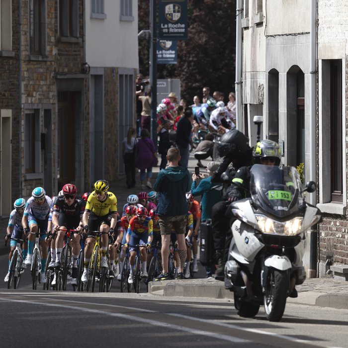 Liege Bastogne Liege 2025 - The peloton approaches up a road lined with tall stone buildings at La Roché-en-Ardenne