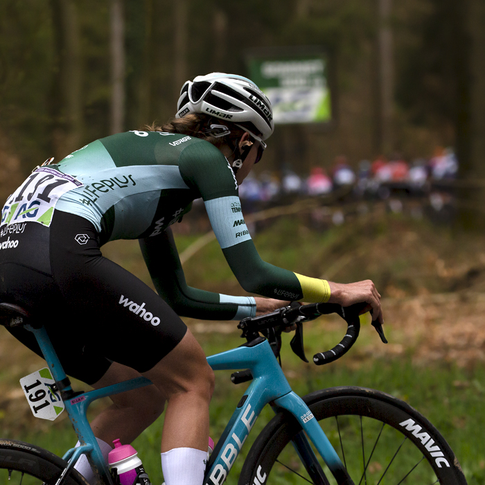 Liège-Bastogne-Liège Femmes 2023 - Lifeplus Wahoo’s Ella Harris climbs through the woodland with the peloton visible through the trees in front