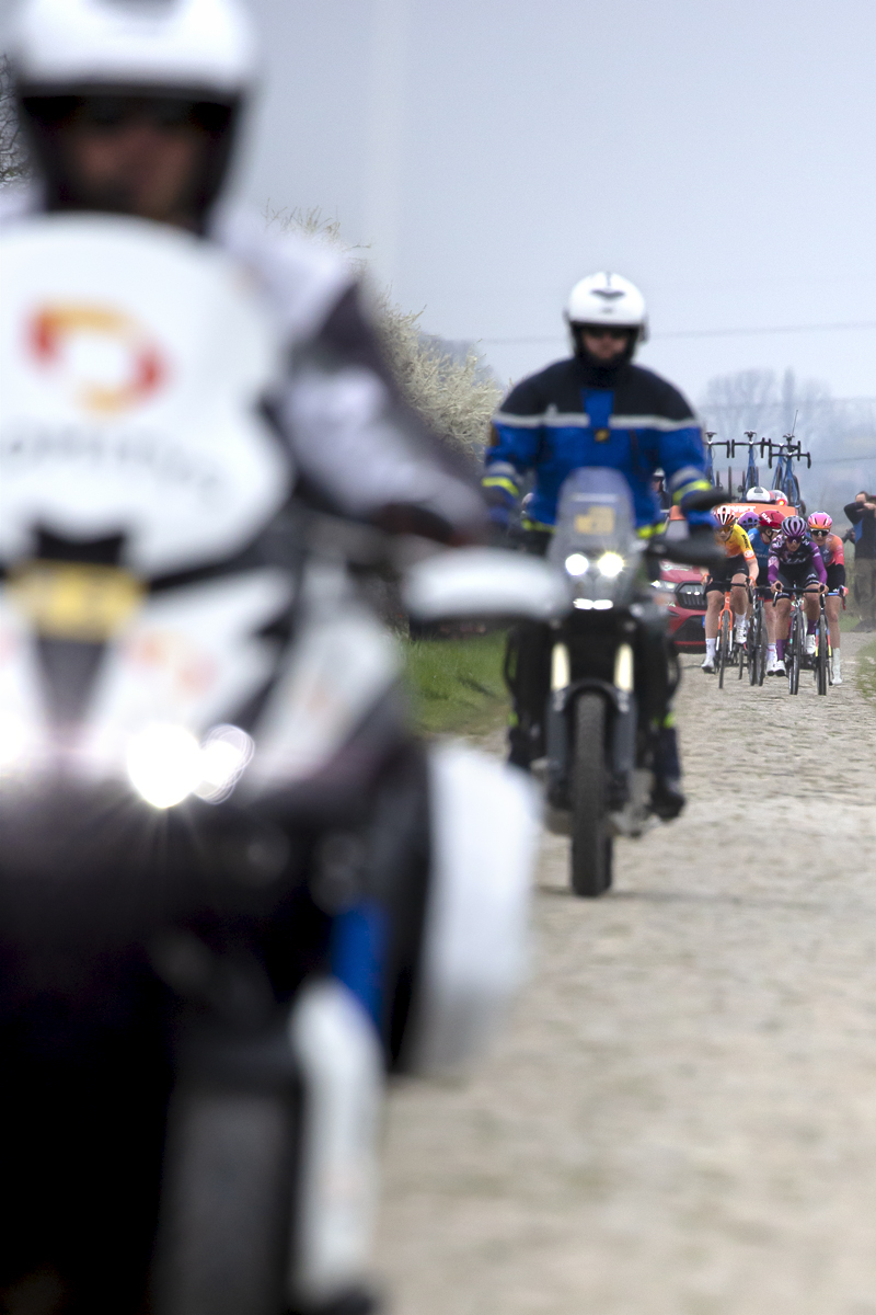 Paris Roubaix Femmes 2023 - Riders approach in the distance on the cobbles while race motorbikes pass in the foreground