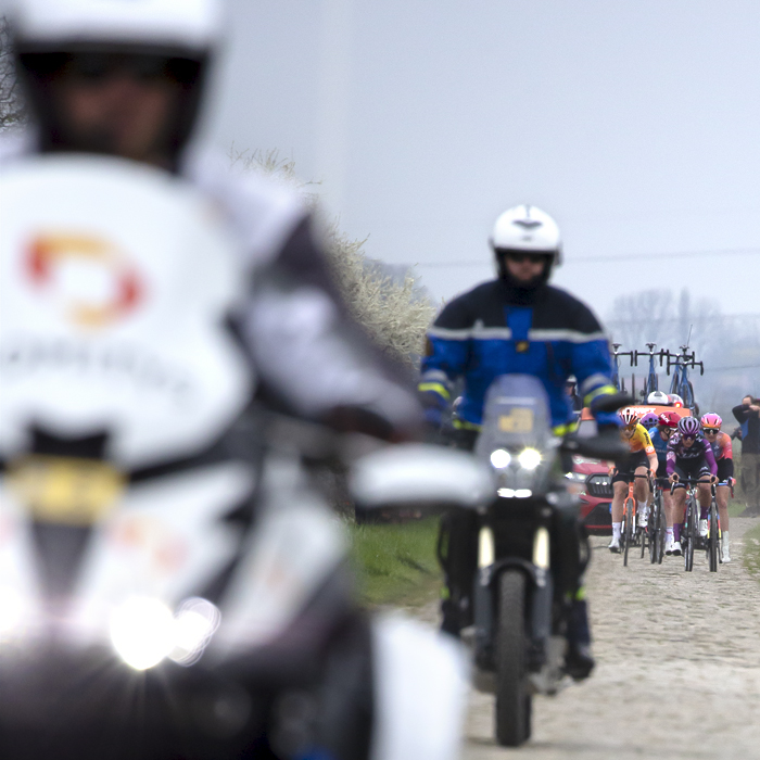 Paris Roubaix Femmes 2023 - Riders approach in the distance on the cobbles while race motorbikes pass in the foreground