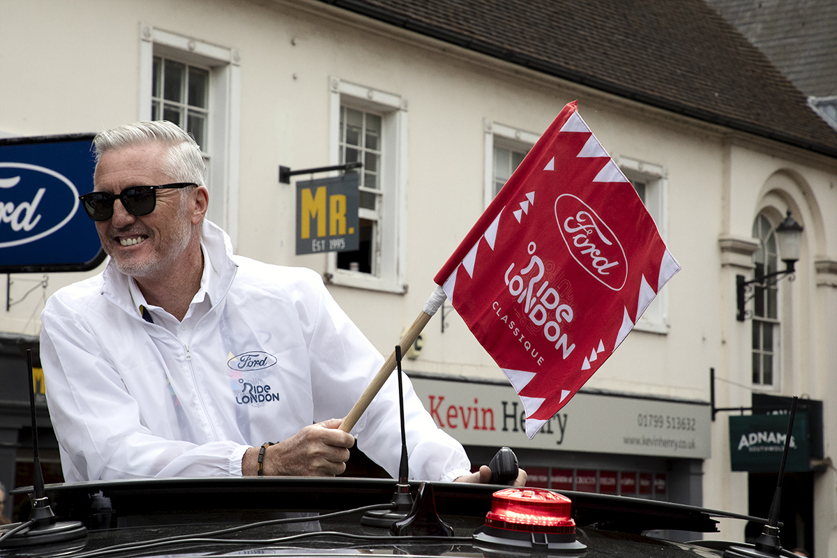 RideLondon Classique 2024 - Race Director, Scott Sunderland holds the race start flag in his hand as he emerges from the sunroof of the race car.