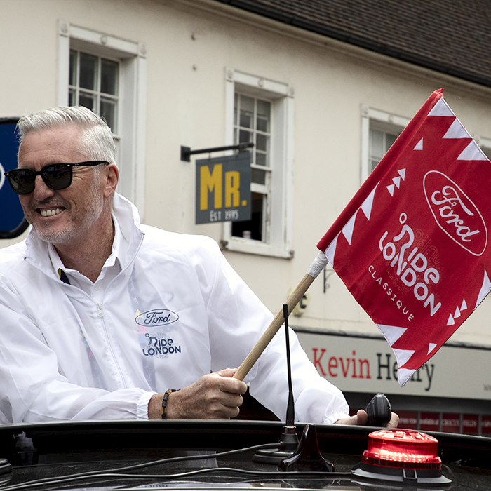 RideLondon Classique 2024 - Race Director, Scott Sunderland holds the race start flag in his hand as he emerges from the sunroof of the race car.