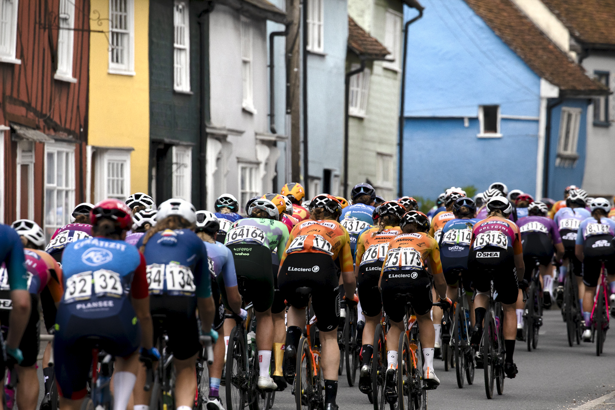 RideLondon Classique 2024 - The peloton moves away past brightly painted cottages