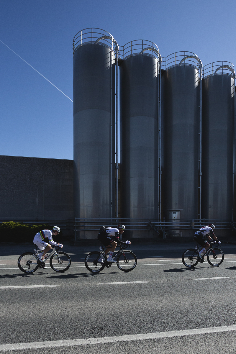 Ronde van Vlaanderen 2025 - A group of elite riders race in front of a line of industrial silos