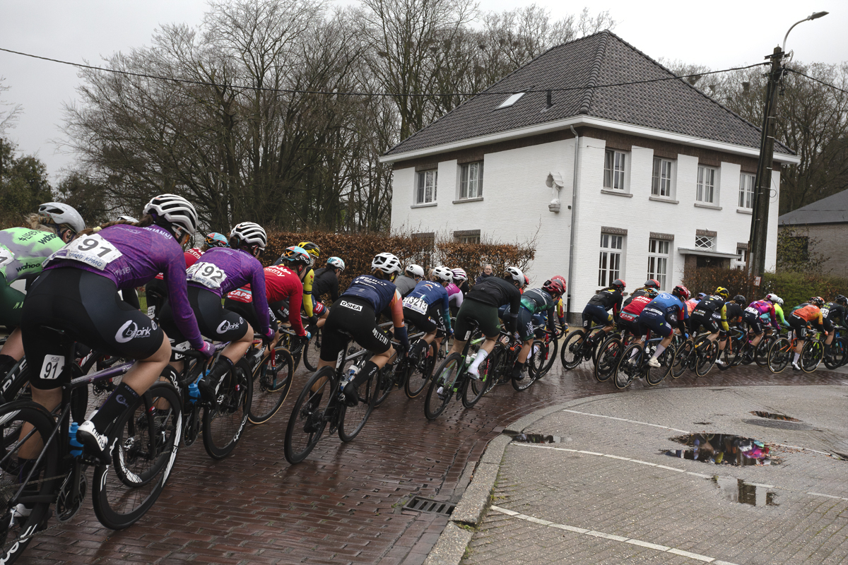 Scheldeprijs Vrouwen 2024 - Riders pass in fort of a white building in Achterbroek
