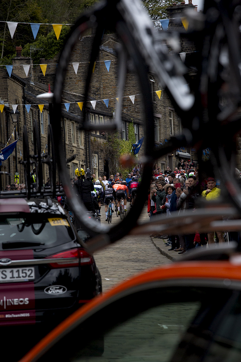 Tour de Yorkshire 2019 - The back of the peloton viewed through a bike wheel on the top of one of the support vehicles, Haworth