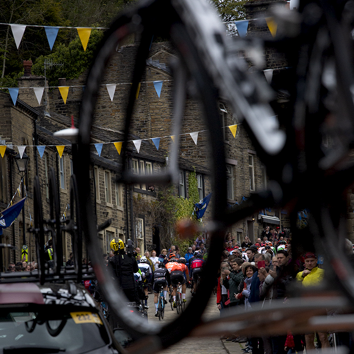 Tour de Yorkshire 2019 - The back of the peloton viewed through a bike wheel on the top of one of the support vehicles, Haworth