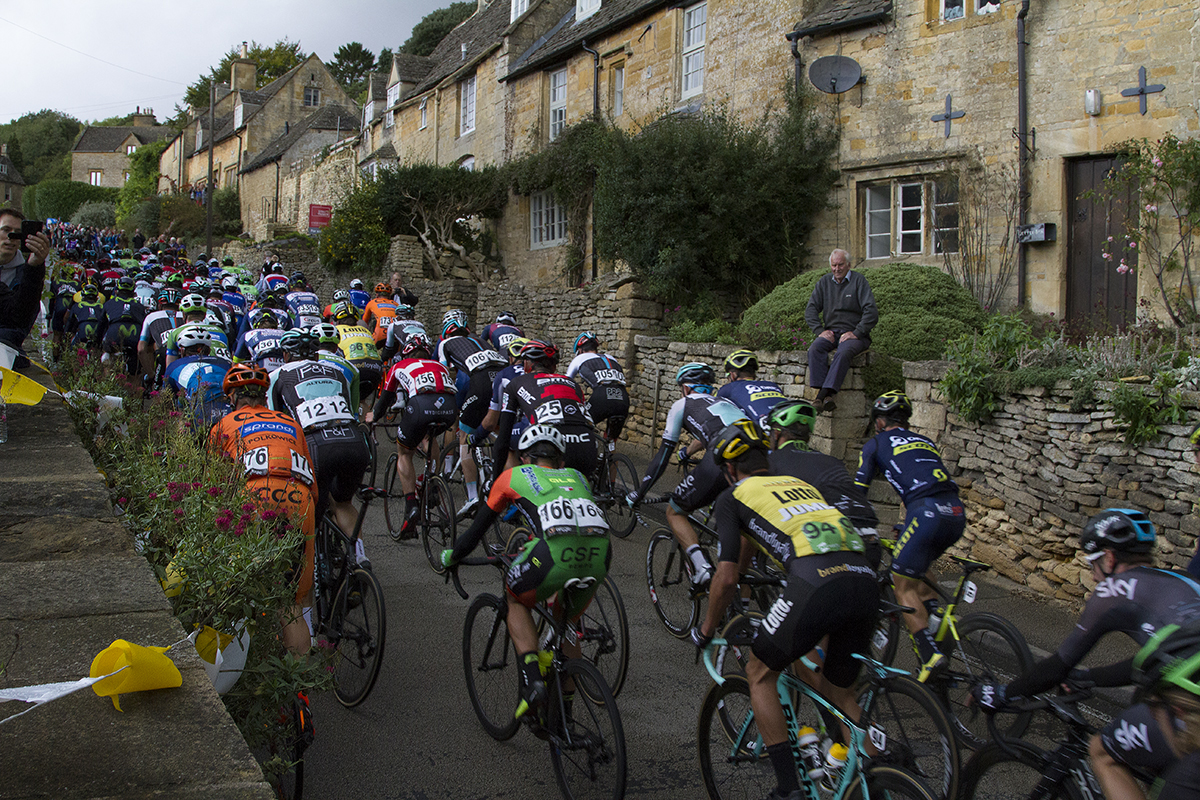 Tour of Britain 2017 - An old man sits on his wall and watches the race as the peloton passes traditional Cotswold cottages