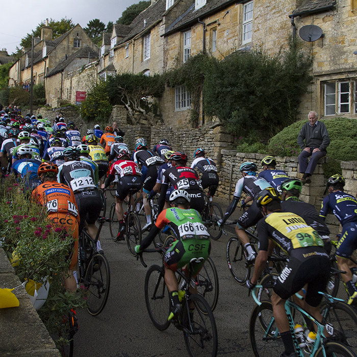 Tour of Britain 2017 - An old man sits on his wall and watches the race as the peloton passes traditional Cotswold cottages