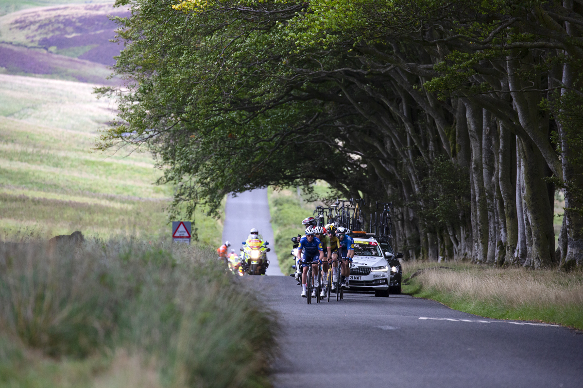 Tour of Britain 2021 - The peloton approaches framed by trees, while heather covered moorland can be seen in the distance