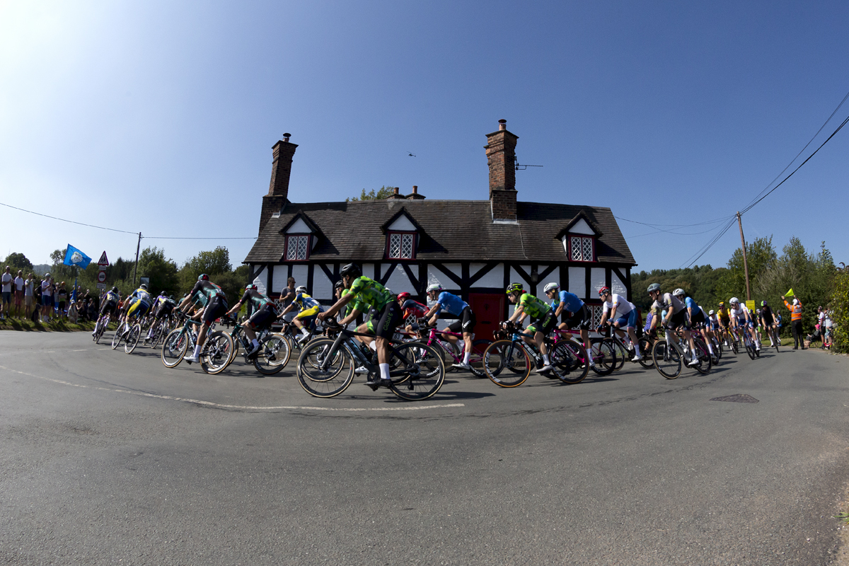 Tour of Britain 2023 - The peloton passes a black and white timber framed cottage on a corner