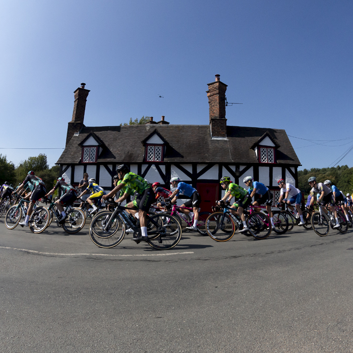 Tour of Britain 2023 - The peloton passes a black and white timber framed cottage on a corner