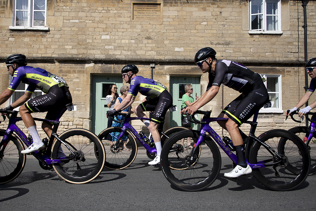 Tour of Britain 2023 - The Bolton Equities Black Spoke team pass by Almshouses in Cirencester as fans look on