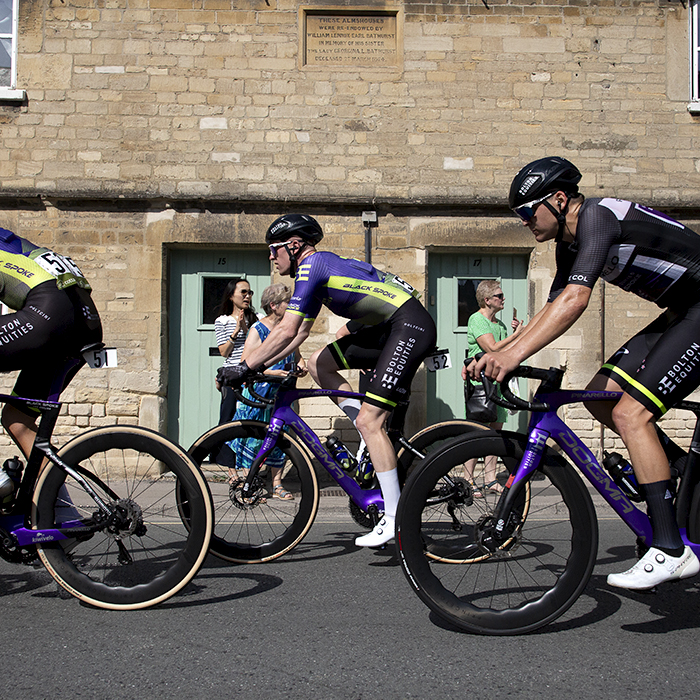 Tour of Britain 2023 - The Bolton Equities Black Spoke team pass by Almshouses in Cirencester as fans look on