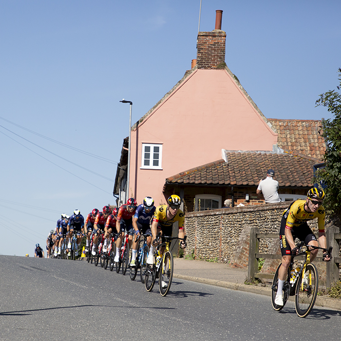 Tour of Britain 2023 - A strung out peloton passes by traditional Suffolk Pink houses in Debenham