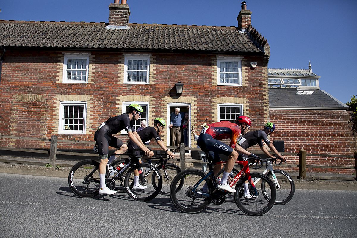 Tour of Britain 2023 - A group of riders pass a brick cottage while elderly fans watch from the doorway