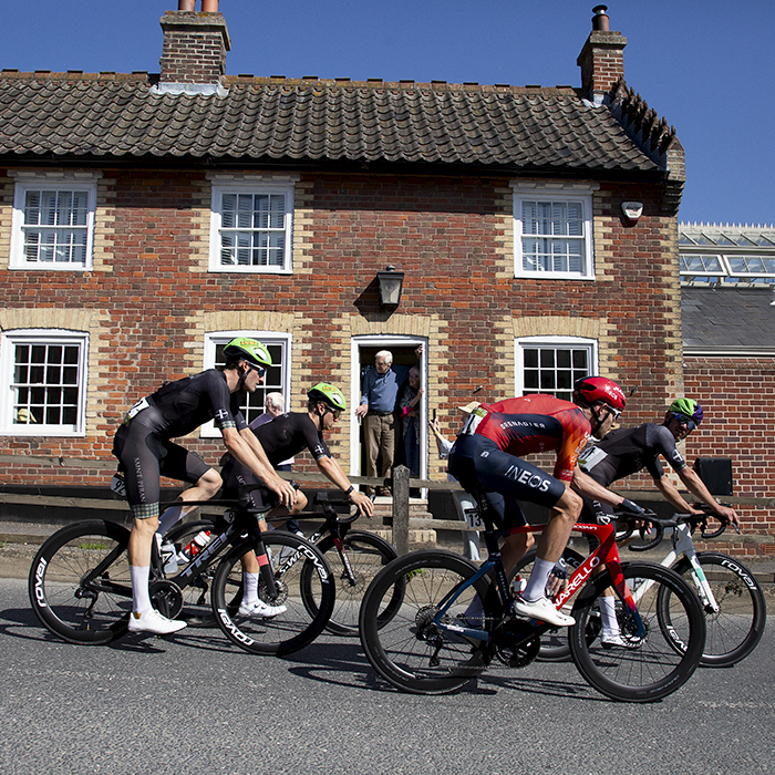 Tour of Britain 2023 - A group of riders pass a brick cottage while elderly fans watch from the doorway