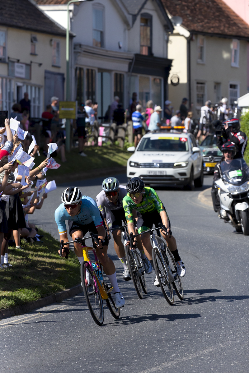 Tour of Britain 2023 - Riders race through Debenham passing a class of school children waving flags they have made