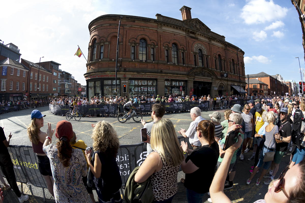 Tour of Britain 2023 - Fans film riders on their phones as they pass a red brick building in Manchester