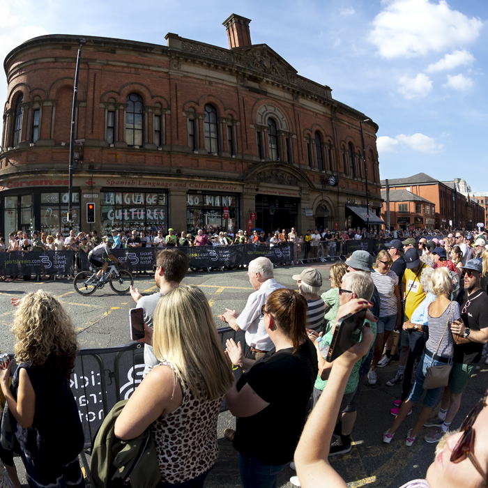 Tour of Britain 2023 - Fans film riders on their phones as they pass a red brick building in Manchester