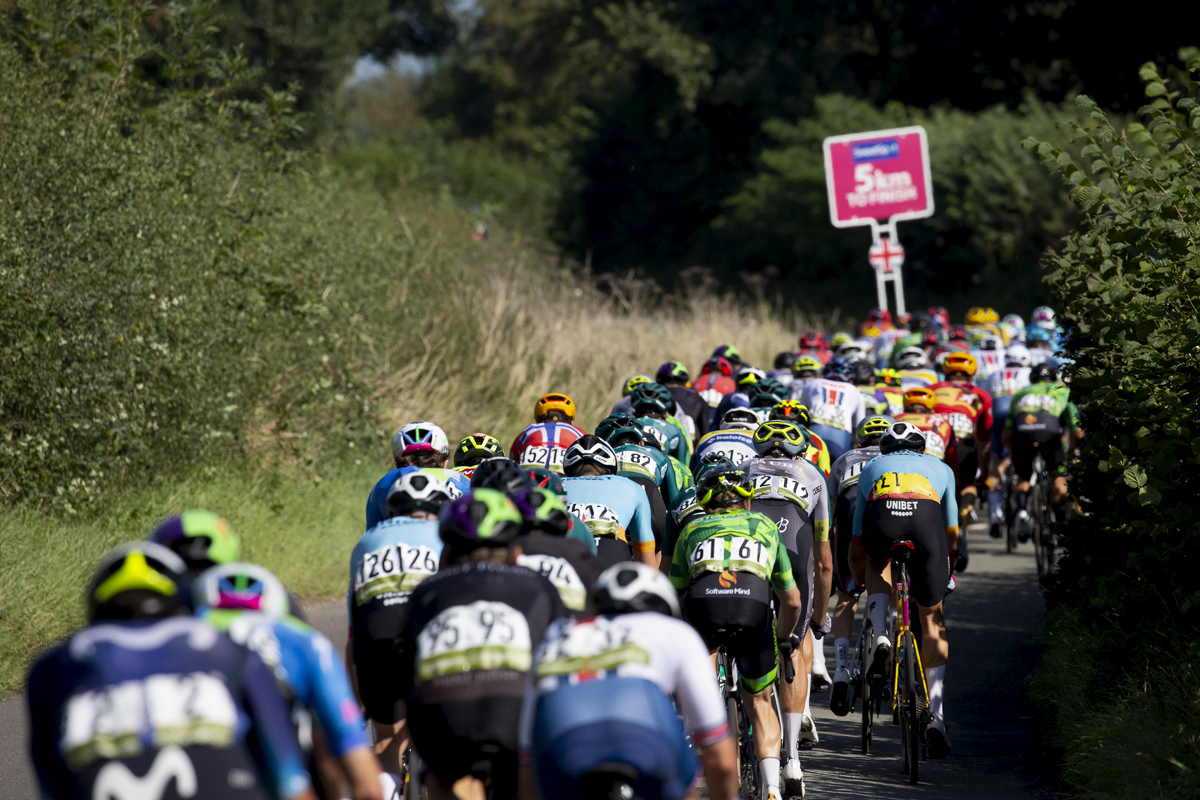 Tour of Britain 2023 - The peloton from the rear as it passes down a narrow hedge lined road with the 5km to go sign in the background