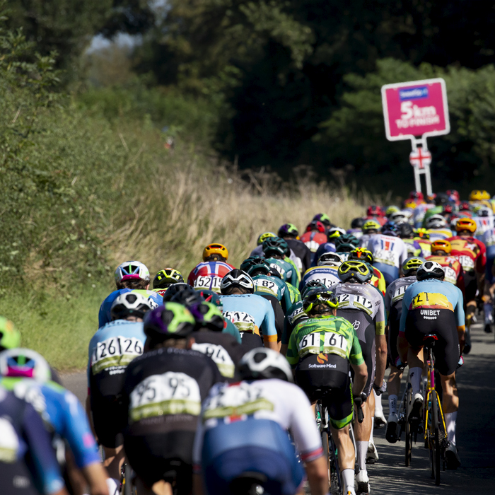 Tour of Britain 2023 - The peloton from the rear as it passes down a narrow hedge lined road with the 5km to go sign in the background