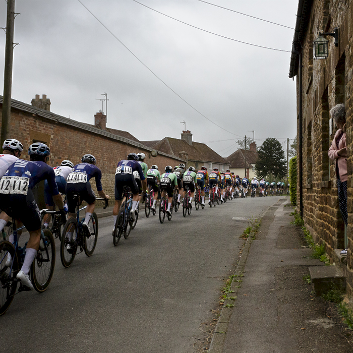 Tour of Britain 2024 - Riders pass a stone built row of cottages as a woman watches on from her doorstep