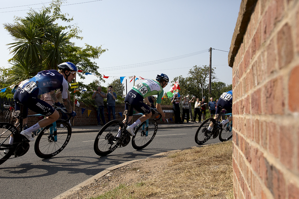 Tour of Britain 2024 - Stevie Williams and his team mates round a corner over a bridge past fans flying a Welsh flag