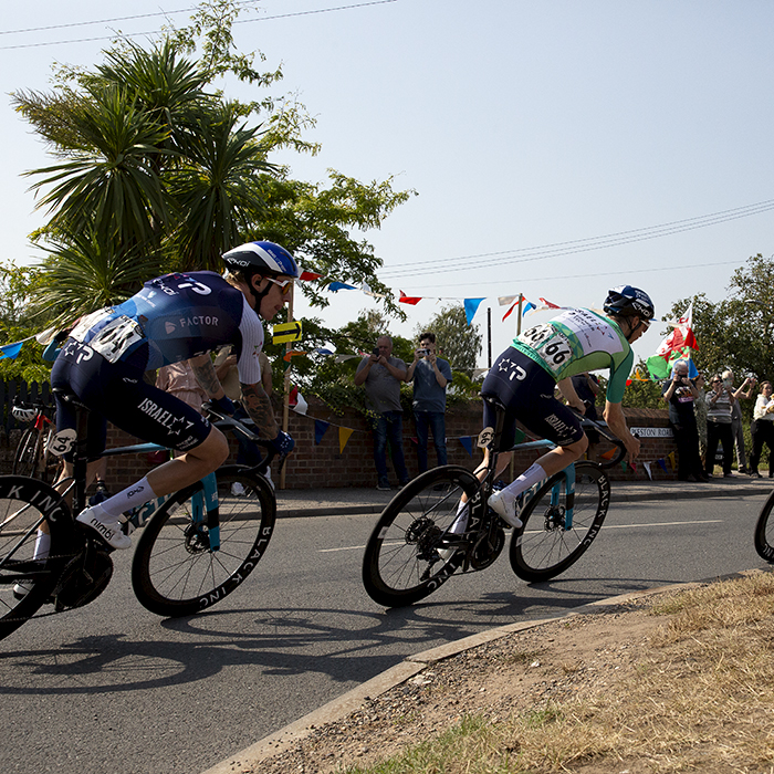 Tour of Britain 2024 - Stevie Williams and his team mates round a corner over a bridge past fans flying a Welsh flag