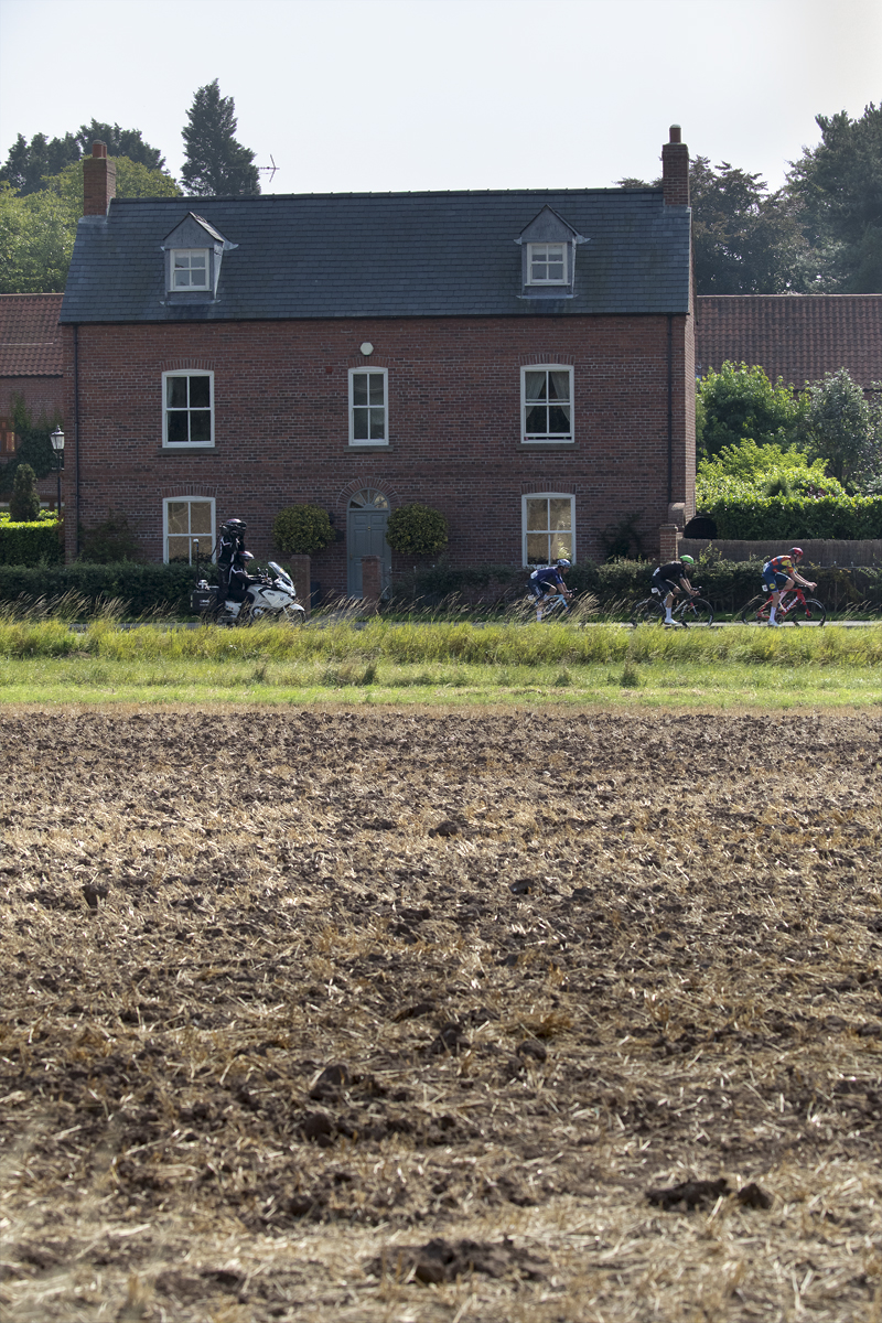 Tour of Britain 2024 - Riders seen over a ploughed field passing by a house on their way into Egmanton