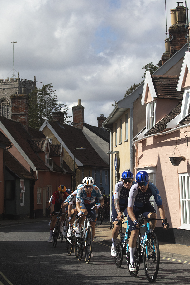 Tour of Britain 2024 - Riders pass through a narrow street of brightly coloured cottages with the village church in the background