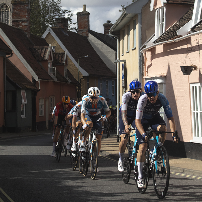 Tour of Britain 2024 - Riders pass through a narrow street of brightly coloured cottages with the village church in the background