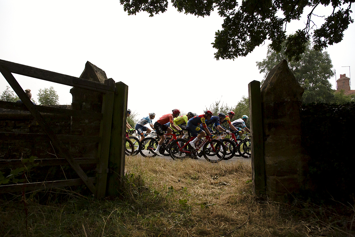 Tour of Britain 2024 - The peloton is framed in the gate of St. John's Church Tower, Little Brington