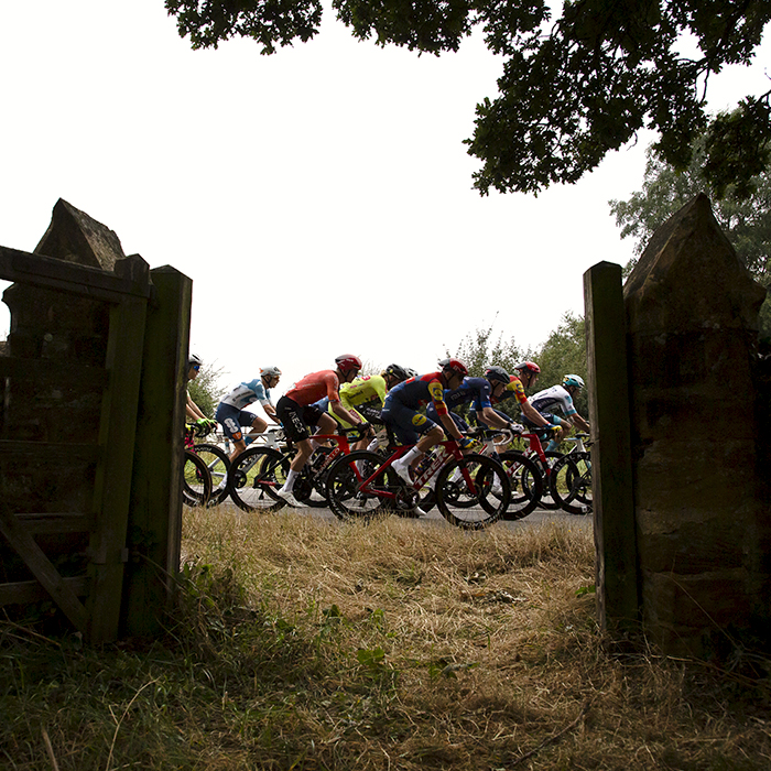 Tour of Britain 2024 - The peloton is framed in the gate of St. John's Church Tower, Little Brington