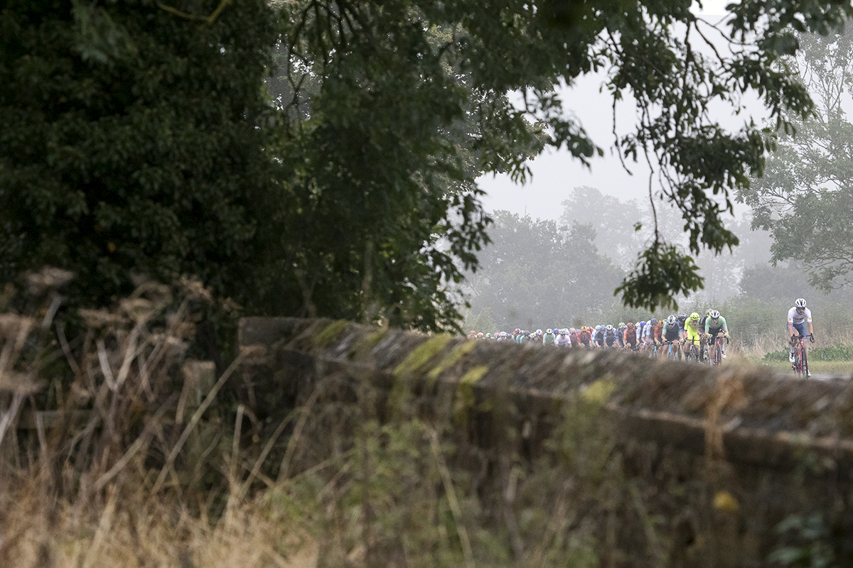 Tour of Britain 2024 - The peloton seen with the stone wall of a churchyard in the foreground