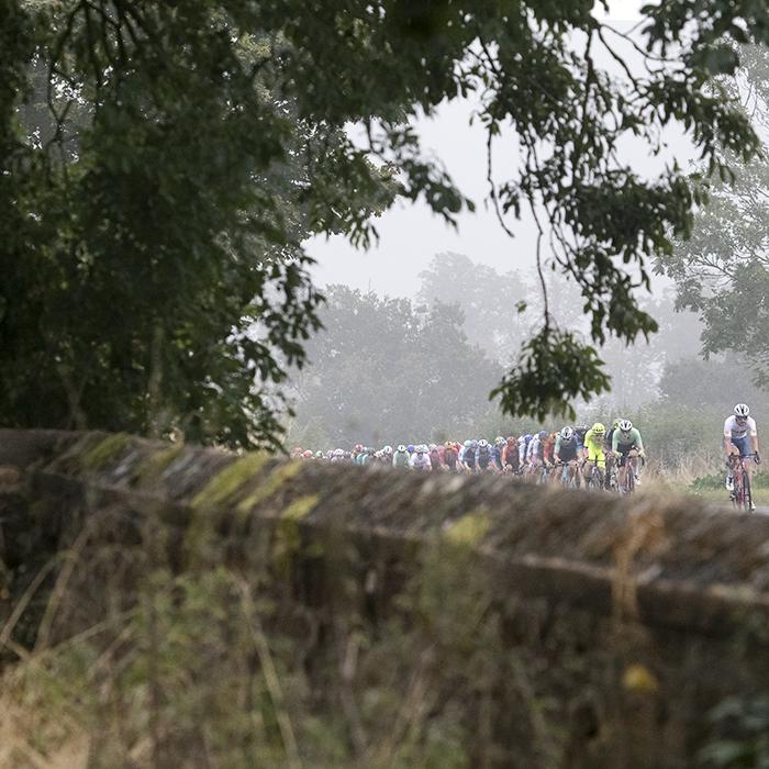 Tour of Britain 2024 - The peloton seen with the stone wall of a churchyard in the foreground